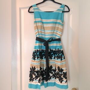 Teal, khaki, and black cocktail dress with black floral design and black belt.